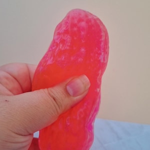 Może przedstawiać: Półprzezroczysta, neonowo-różowa, miękka zabawka sensoryczna o wyboistej, teksturowanej powierzchni. Materiał jest elastyczny i podatny na formowanie, wykazując żywy, półprzezroczysty blask, gdy światło przenika przez żelową substancję.