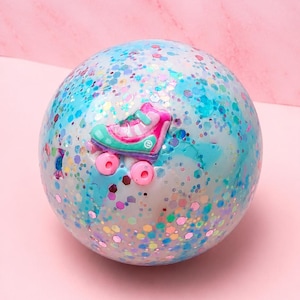 May include: A spherical bath bomb with a white base, marbled with light blue swirls and embedded with multicoloured hexagonal glitter. A small, three-dimensional pink and teal roller skate charm is attached to the centre of the sphere.