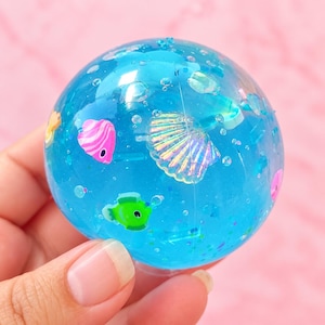 May include: A clear blue spherical stress ball filled with liquid, containing small floating plastic figures including a pink fish, a green fish, and an iridescent seashell, along with tiny glitter particles suspended throughout the blue gel.