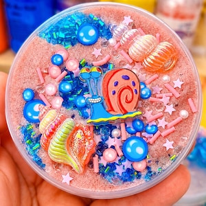May include: A round container filled with pink textured slime, featuring a blue snail character charm, two iridescent seashell charms, blue and pink beads, small pink star-shaped sprinkles, and blue glitter accents scattered throughout the surface.