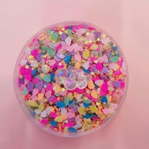 May include: A clear plastic container filled with colorful confetti and glitter. The confetti includes star and cloud shapes in pink, yellow, blue, and purple. A clear, bubble-shaped decoration sits in the center, all on a pink background.
