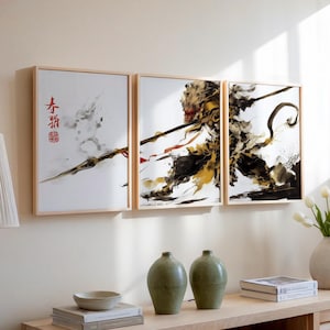 Può includere: Tre stampe d'arte incorniciate che raffigurano un'illustrazione stilizzata di un guerriero scimmia che tiene una lancia. La stampa più a sinistra presenta caratteri cinesi in inchiostro rosso e nero. Le stampe centrale e destra presentano il guerriero scimmia in inchiostro nero, bianco e oro.