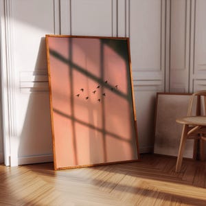 Könnte beinhalten: Ein rechteckiger Wandkunstdruck in einem Holzrahmen mit einem weichen pfirsich- und orangefarbenen Verlaufs-Hintergrund und einem Schwarm kleiner schwarzer Vögel, die durch die Mitte fliegen. Der Druck zeigt ein gitterartiges Schattenmuster.