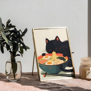 May include: A gold-framed picture of a black cat eating ramen noodles. The picture is on a gold easel and sitting on a pink surface. There is a clear glass vase with green leaves and a brown candle on the surface.