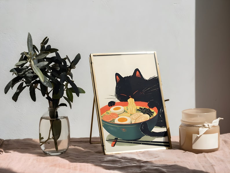May include: A gold-framed picture of a black cat eating ramen noodles. The picture is on a gold easel and sitting on a pink surface. There is a clear glass vase with green leaves and a brown candle on the surface.