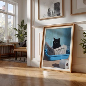 May include: Framed art print featuring a black cat sitting in a blue litter box and reading a newspaper titled "The daily news." A roll of toilet paper is next to the litter box. The artwork is in a light wood frame.