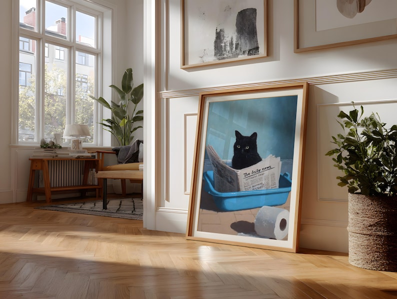 May include: Framed art print featuring a black cat sitting in a blue litter box and reading a newspaper titled "The daily news." A roll of toilet paper is next to the litter box. The artwork is in a light wood frame.