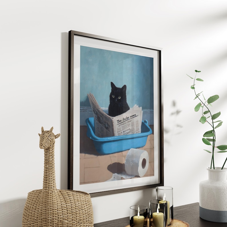 May include: Framed art print featuring a black cat sitting in a blue litter box and reading a newspaper titled "The daily news." A roll of toilet paper sits on the floor. The artwork is displayed on a white wall.
