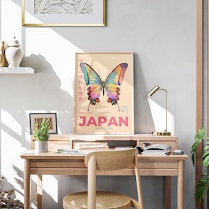 May include: A vintage poster with a colorful butterfly illustration and the word "JAPAN" in pink letters. The poster is framed and hanging on a wall above a wooden desk with a lamp, books, and a plant.