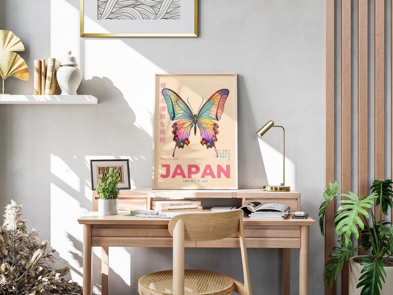 May include: A vintage poster with a colorful butterfly illustration and the word "JAPAN" in pink letters. The poster is framed and hanging on a wall above a wooden desk with a lamp, books, and a plant.
