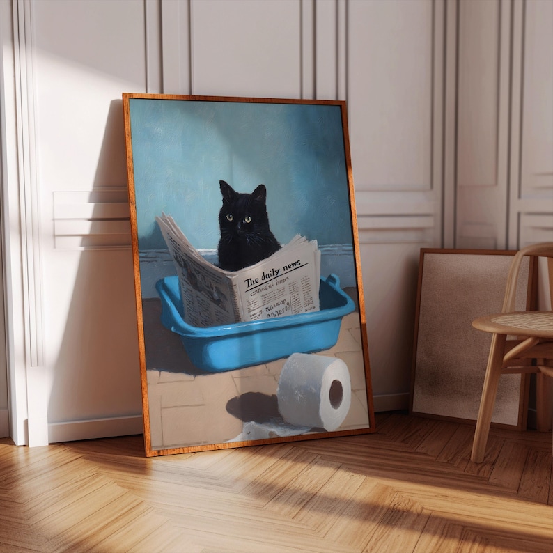 May include: Framed art print featuring a black cat sitting in a blue litter box, reading a newspaper titled "The daily news." A roll of toilet paper sits nearby. The artwork has a wooden frame and is displayed on a wooden floor.