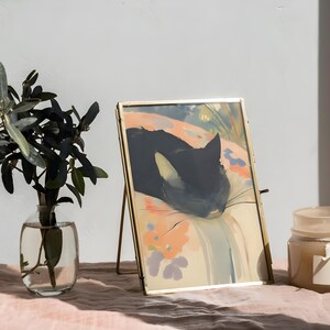 Puede incluir: Una imagen enmarcada en oro de un gato negro durmiendo sobre un fondo colorido con estampado floral. La imagen est&aacute; sobre un caballete dorado y se encuentra sobre una mesa rosa.
