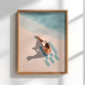 May include: A framed art print depicts a person reading on a beach towel. The artwork features a person in a white swimsuit, a blue and white striped towel, and a light blue sky. The print is in a wooden frame.