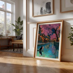 May include: A framed art print depicts a nighttime scene with a river, buildings, and cherry blossom trees. A black cat is silhouetted on a small boat. The artwork is in a light wood frame, leaning against a white wall.