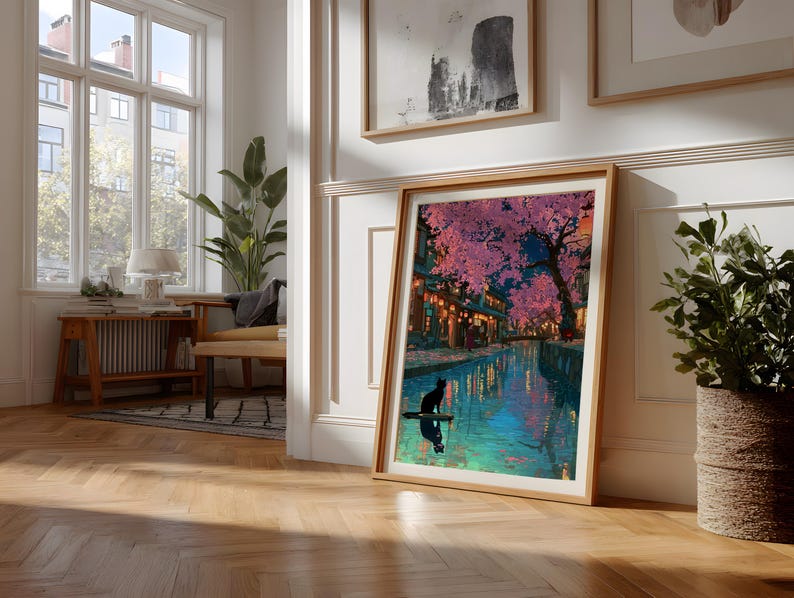 May include: A framed art print depicts a nighttime scene with a river, buildings, and cherry blossom trees. A black cat is silhouetted on a small boat. The artwork is in a light wood frame, leaning against a white wall.