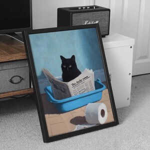 May include: Framed art print featuring a black cat sitting in a blue litter box, reading a newspaper titled "The Daily News". A roll of toilet paper sits nearby. The background is a light blue wall.