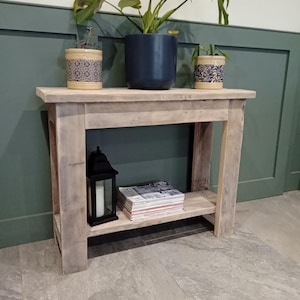 May include: A rustic wooden console table with a shelf, featuring a distressed white finish. The table is decorated with three potted plants, a black lantern, and a stack of magazines.