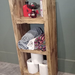 Puede incluir: Estantería de madera rústica con tres estantes. El estante superior tiene una botella roja y una botella rosa con un diseño de flor negra. El estante del medio tiene tres toallas enrolladas, una gris, una blanca y una con rayas de colores. El estante inferior tiene tres rollos de papel higiénico.
