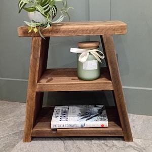 May include: A brown wooden two-tier side table with angled legs. The top surface holds a small potted green plant. The middle shelf holds a light green candle in a glass jar with a wooden lid and a white ribbon. A book titled The Front is on the bottom.