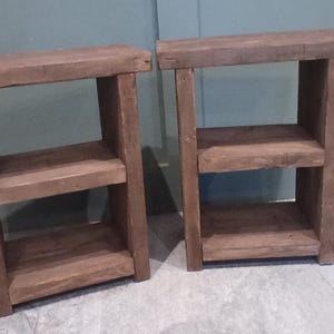 May include: Two rustic, brown wooden side tables. Each table has a rectangular top, a middle shelf, and a bottom shelf. The tables are constructed with visible wood grain and a simple, sturdy design. Ideal for a living room or bedroom.