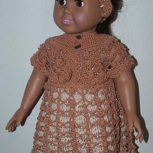 May include: A brown crocheted dress with a white crocheted headband. The dress has a scalloped hem and a button closure at the neckline. The doll is wearing the dress and headband.