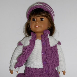 Puede incluir: Una muñeca con un atuendo de ganchillo morado y blanco. El atuendo incluye un sombrero, un chaleco, una camisa de manga larga y una falda. La muñeca tiene el pelo castaño y los ojos marrones.