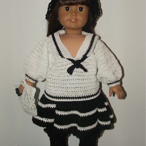 Puede incluir: Una muñeca con un traje de marinero de ganchillo blanco y negro con una boina negra. La muñeca tiene el pelo castaño y los ojos marrones. La muñeca está de pie sobre una superficie de mimbre.