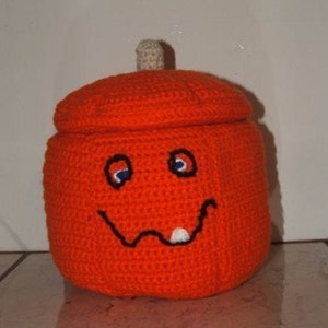May include: A crocheted orange pumpkin with a smiling face, two eyes, and a single tooth. The pumpkin has a lid and is perfect for holding small items.