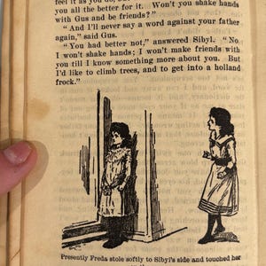 K&ouml;nnte beinhalten: Schwarz-wei&szlig;-Illustration von zwei M&auml;dchen, die in einem T&uuml;rrahmen stehen. Das M&auml;dchen links tr&auml;gt ein langes Kleid und das M&auml;dchen rechts tr&auml;gt ein Kleid mit Matrosenkragen. Der Text "Presently Freda stole softly to Sibyl's side and touched her on the arm." ist unter der Illustration gedruckt.