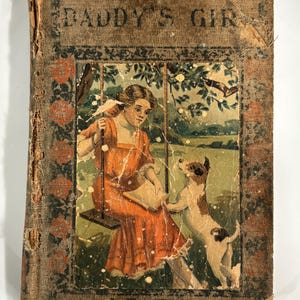 K&ouml;nnte beinhalten: Ein altes Buch mit dem Titel "Daddy's Girl" mit einem abgenutzten braunen Einband. Der Einband zeigt eine Illustration eines M&auml;dchens in einem roten Kleid, das auf einer Schaukel sitzt, mit einem Hund an ihrer Seite. Das Buch ist von einem dekorativen Rahmen umgeben.