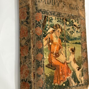 K&ouml;nnte beinhalten: Ein altes Buch mit dem Titel "Daddy's Girl" mit einem abgenutzten braunen Einband und einer verblassten Illustration einer Frau auf einer Schaukel mit einem Hund. Das Buch hat ein Blumenmuster auf dem Einband.