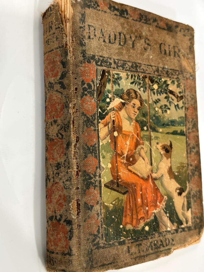 K&ouml;nnte beinhalten: Ein altes Buch mit dem Titel "Daddy's Girl" mit einem abgenutzten braunen Einband und einer verblassten Illustration einer Frau auf einer Schaukel mit einem Hund. Das Buch hat ein Blumenmuster auf dem Einband.