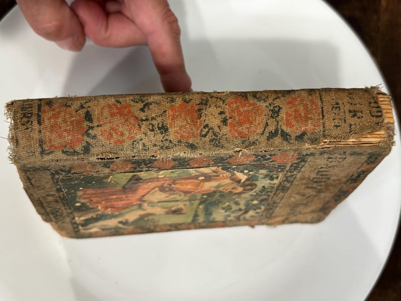K&ouml;nnte beinhalten: Ein altes Buch mit einem abgenutzten braunen Einband und einer verblassten Illustration einer Frau in einem roten Kleid. Der Buchr&uuml;cken hat japanischen Text und die Worte "Die kleine rote Henne" stehen auf dem Einband.