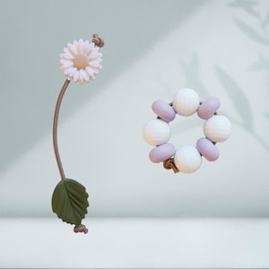 May include: A white daisy with a brown stem and green leaf attached to a purple and white beaded bracelet. The bracelet is made of silicone beads and has a knotted design.