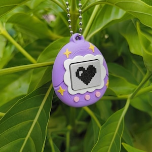 May include: A purple digital pet toy with a pixelated black heart on the screen. The toy has yellow star and pink dot accents, and hangs from a silver ball chain. The background is lush green foliage.