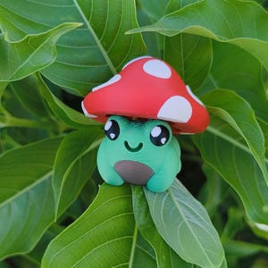 Könnte beinhalten: Eine skurrile, 3D-gedruckte Figur einer grünen Frosch mit einem roten Pilzhut mit weißen Punkten. Der Frosch hat große, ausdrucksstarke Augen und einen lächelnden Mund. Die Figur ist vor dem Hintergrund üppiger grüner Blätter platziert.