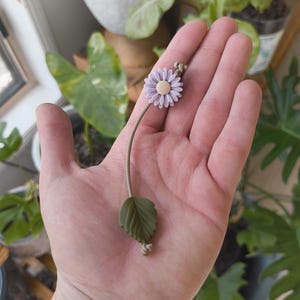 Op de afbeelding: Een delicate paarse bloem met een enkel groen blad aan een bruin touwtje. De bloem is gemaakt van polymer clay en is een handgemaakte charme.