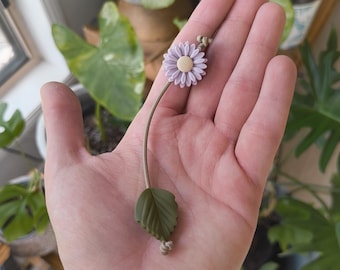 May include: A delicate purple flower with a single green leaf on a brown string. The flower is made of polymer clay and is a handmade charm.