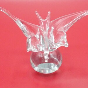 May include: A clear glass sculpture of a butterfly with its wings spread wide. The butterfly is perched on a round, clear glass base.