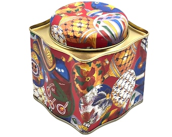 May include: A decorative, hexagonal tin with a removable lid. The tin and lid are red with a repeating pattern of colorful hot air balloons. The tin has a gold-colored rim and a gold-colored interior.