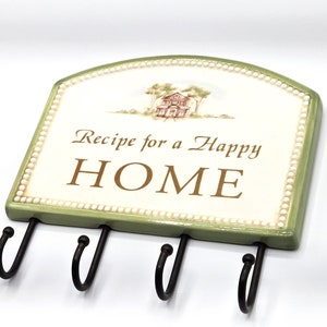 May include: A green and white ceramic key holder with a white background and a green border. The key holder has a white house illustration and the text "Recipe for a Happy HOME". The key holder has four black metal hooks.