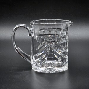 May include: A clear glass pitcher with a cut glass design. The pitcher has a handle and a spout.