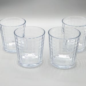 May include: Four clear, cylindrical drinking glasses. Each glass has a textured, grid-like pattern on the exterior. The glasses are empty and appear to be made of glass or a similar transparent material. The glasses are arranged on a white surface.
