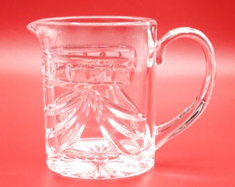 Könnte beinhalten: Ein Sahnekännchen aus klarem Glas mit facettiertem Schliffmuster, vertikalen Linien und einem Sternmuster am Boden. Das Kännchen verfügt über einen geschwungenen Henkel auf der rechten Seite und einen kleinen Ausguss auf der linken Seite.