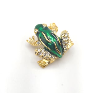 Puede incluir: Broche de rana dorada con cuerpo de esmalte verde y detalles de cristal transparente. Los ojos de la rana también están adornados con cristales. El broche está sobre un fondo blanco.