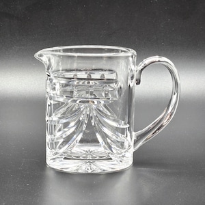 May include: A clear glass pitcher with a cut glass design. The pitcher has a handle and a wide mouth.
