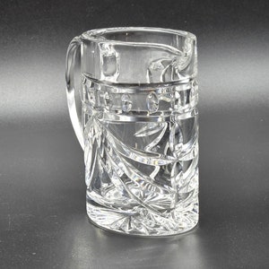 May include: A clear crystal pitcher with a cut glass design. The pitcher has a handle and a wide mouth.