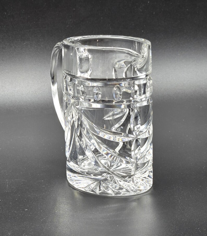 May include: A clear crystal pitcher with a cut glass design. The pitcher has a handle and a wide mouth.