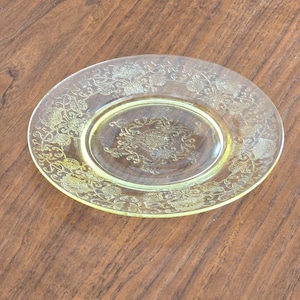 Può includere: Un piatto di vetro trasparente tinto di giallo con un intricato disegno floreale inciso sul bordo e al centro. Il piatto è rotondo, con un bordo leggermente rialzato, e poggia su una superficie di legno. Il diametro del piatto è di circa 20 cm.