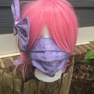 May include: A pink wig with a purple and white bow with a syringe and bandage pattern. The wig is wearing a purple face mask with a syringe and bandage pattern.